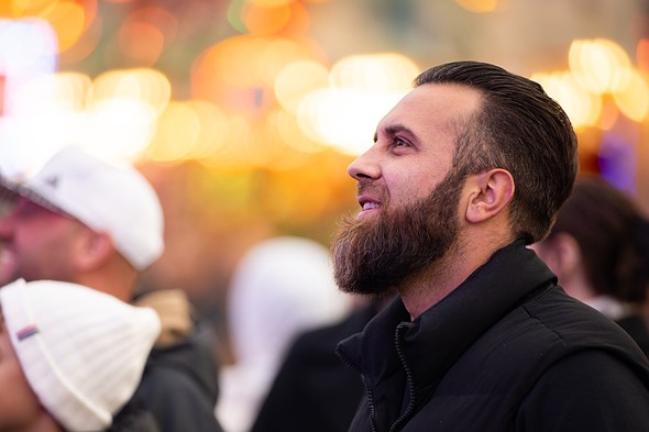 a bearded man with a black coat stands looking ahead while smiling in front of blurred lights and people