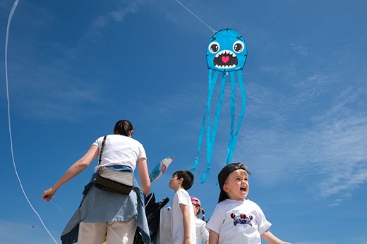 a blue kite with a silly face flies  over a group and child making a similar face