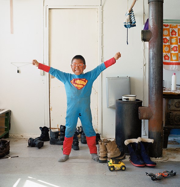 a boy in a worn superman costume and orange goggles stands smiling with arms up in a room with winter boots2