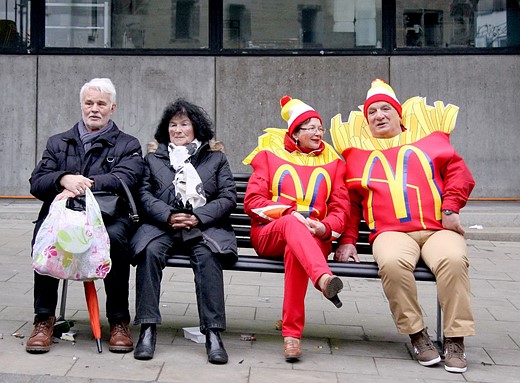 a couple in mcdonalds frie costumes sits on a bench next to a couple in black coats