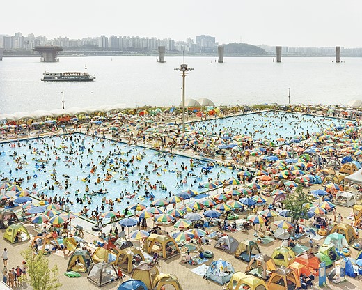 a crowd of people swims at two pools on the shore of a body of water with a cityscape in the background