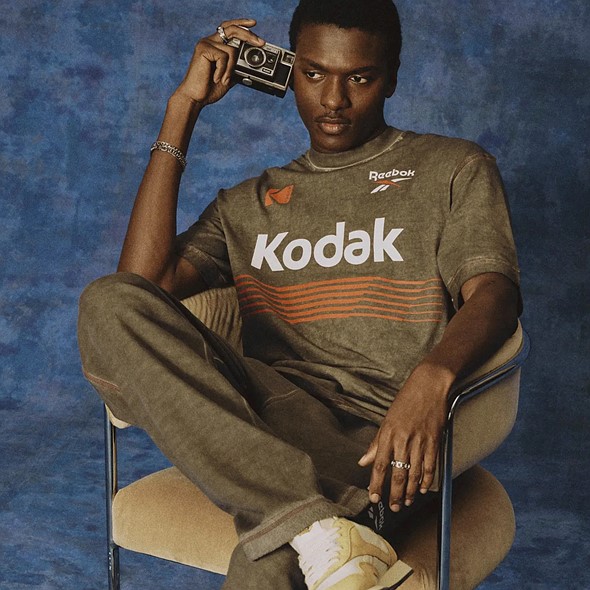 a man lounges in a chair with is leg up while holding an old camera and wearing a shirt with kodak and reebok logos
