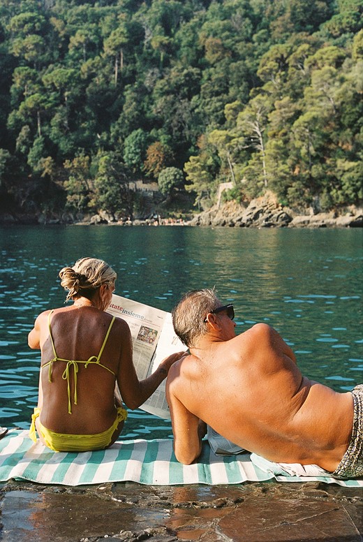a man lounges on a towel next to a woman reading a newspaper in front of a lake