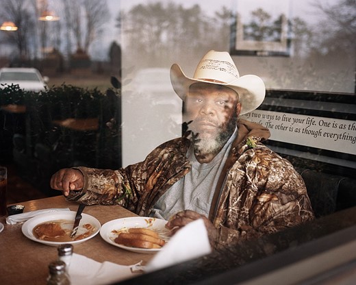 a man sits at a dine booth with plates of food and reflections on a window in front of Him