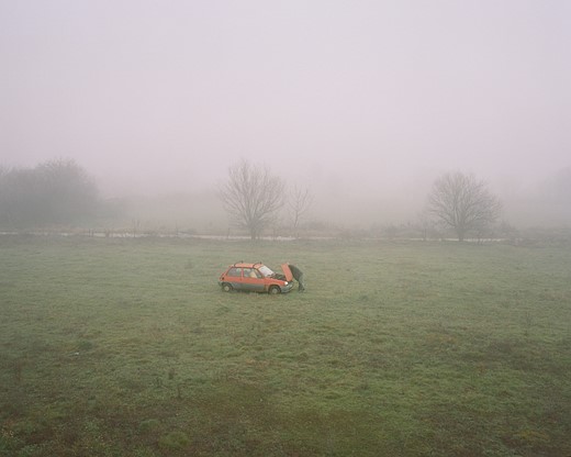 a man stands over the open hood of a red car in a foggy field