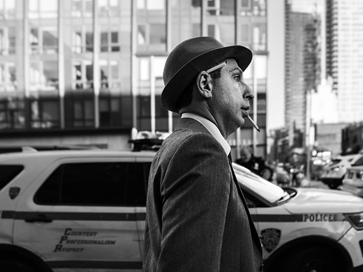 a man wearing a hat has a cigarette in his mouth and behind his year walks down a street