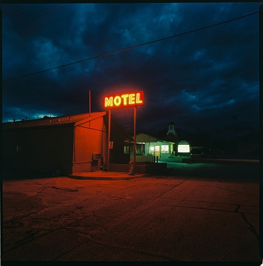 a neon motel sign is lit up in front of a building with dark clousd looming overhead