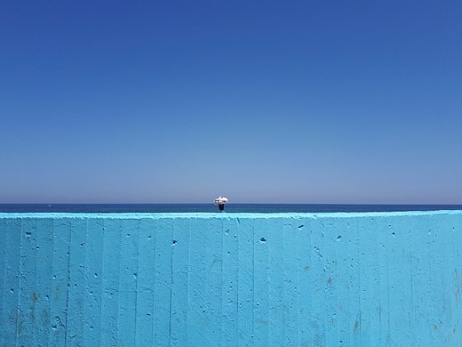 a person carrying a white bundle on their head walks behind a blue wall with water and blue sky behind