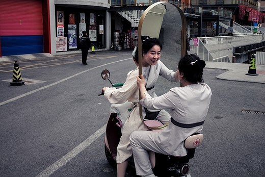 a person rides a motorbike while holding a large mirror reflecting her smiling face