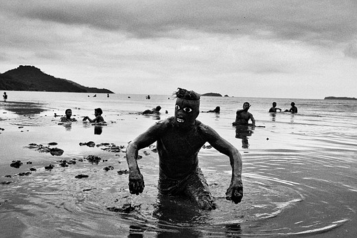a person wearing a mask with large painted eyes comes out of the water with people behind hime