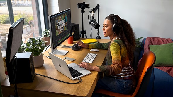 a woman sits at a desk with a podcasting setup including a camera mic and multiple computers