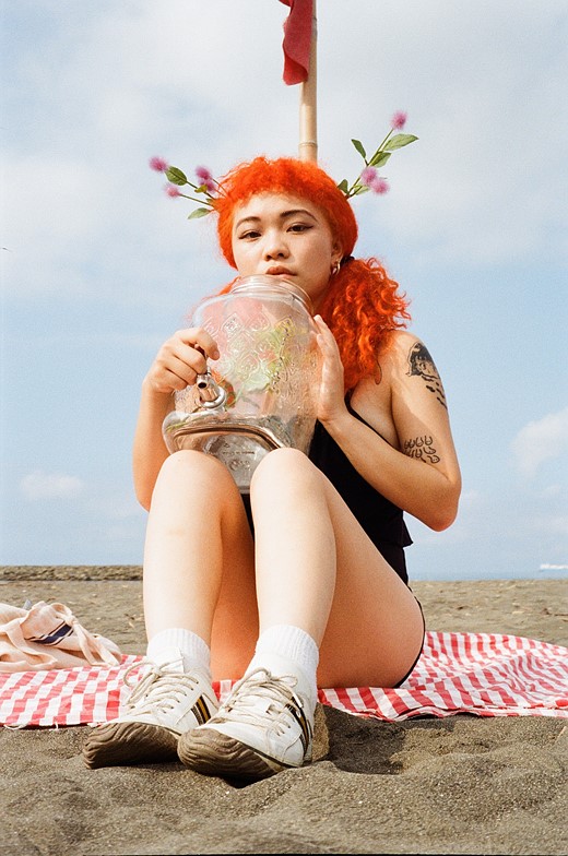 a woman with bright orange hear sits on a red striped towel on a beach while holding a large glass jug