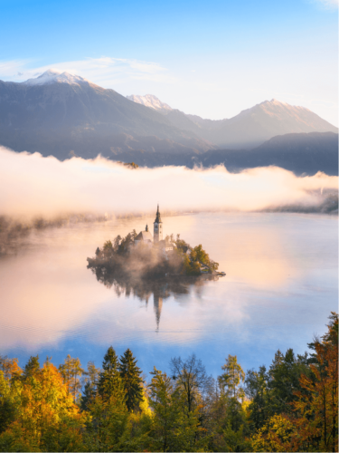 Morning fog surrounding a small island church on a calm lake, illuminated by gentle transition light reflecting off the water and mist.