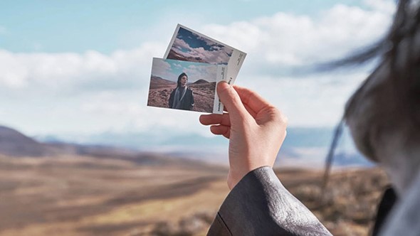 a hand holds two photo prints in front of a vast open landscape