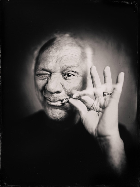 a man squinting one eye holds something to his mouth between his thumb and pointer finger-