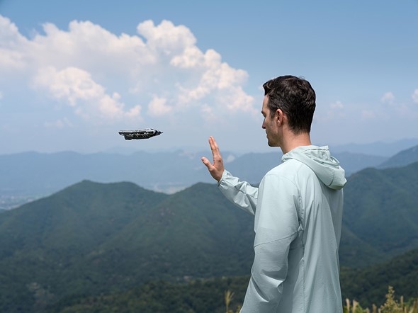 a man stands with his hand up while a drone hovers in front and mountains extend in the background