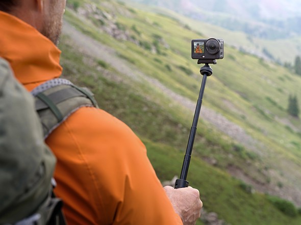 a person in an orange jacket holds a selfi stick with dji osmo action 6 while in the mountains