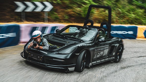 a person wearing a helmet and holding a camera sits in the front trunk of a porsche boxster as it speeds down a track