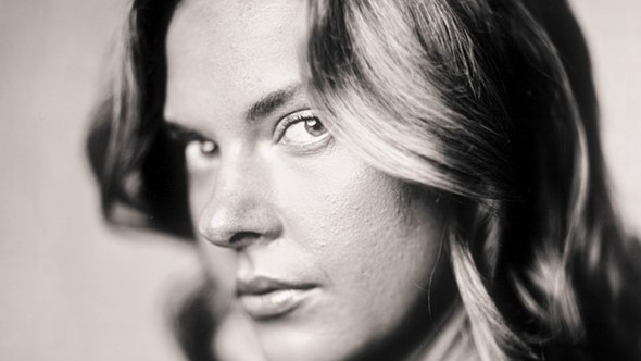 a_woman_with_curled_hair_glances_sideways_while_on_front_of_a_white_background