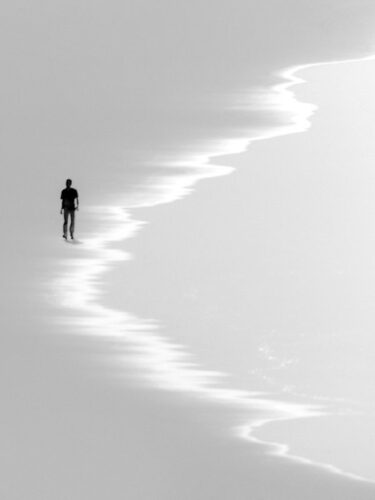 A solitary person walks along a glowing shoreline in soft monochrome light, composing for mood with minimalism and gentle tonal contrast.