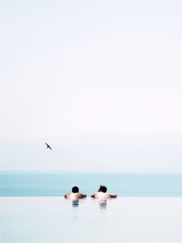 Two swimmers lean on the edge of an infinity pool as a bird flies overhead, using framing techniques to emphasize negative space and serenity.