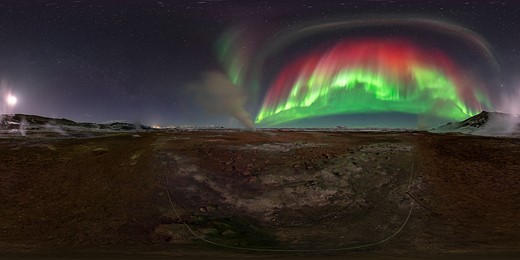 green and red northern lights stretch above a barren rocky valley with snowy mountains on the sides