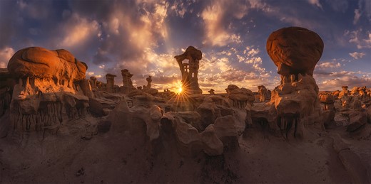 lumpy red rock formations fill a landscape with the sun bursting into view behind one