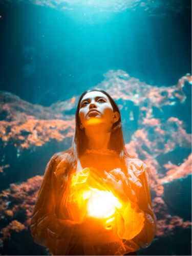 A woman illuminated by a glowing orb held to her chest, surrounded by deep blue underwater tones, showcasing mood editing techniques that blend warm and cool light for dramatic contrast.