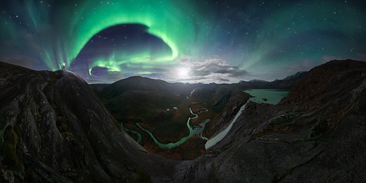 northern lights swirl above a mountainous landscape with a river winding through a canyon
