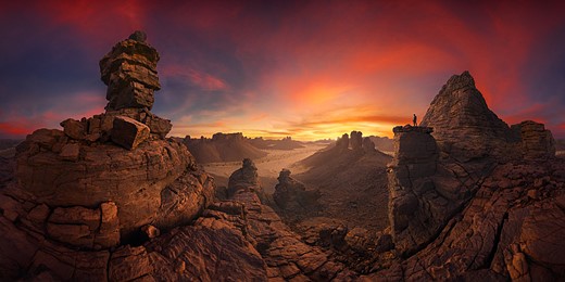 red and orange clouds at Sunset swirl above a lanscape with large red rock formations while a person stands on one pillar