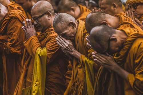 Documentary photo by Marco Tagliarino showing a group of Buddhist monks in saffron robes praying with bowed heads and folded hands.