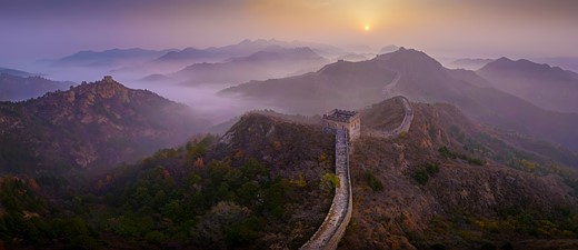 the great wall of china extends down a mountain ridge with fog layered over the mountains in the distance