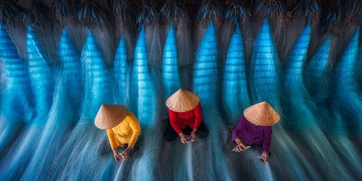 three people wearing straw hats and yellow red and purple shirts sit amidst a blue fishing net