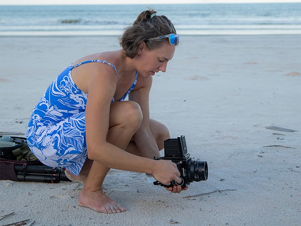 a woman in a blue dress squats down low at the beach while holding a large camera