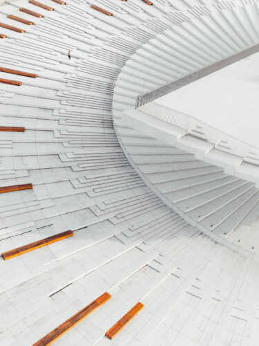 Curved architectural structure with repeating white geometric panels and subtle orange accents, photographed from an unusual perspective as part of Questmas 2025 by Vitaly Tyuk.