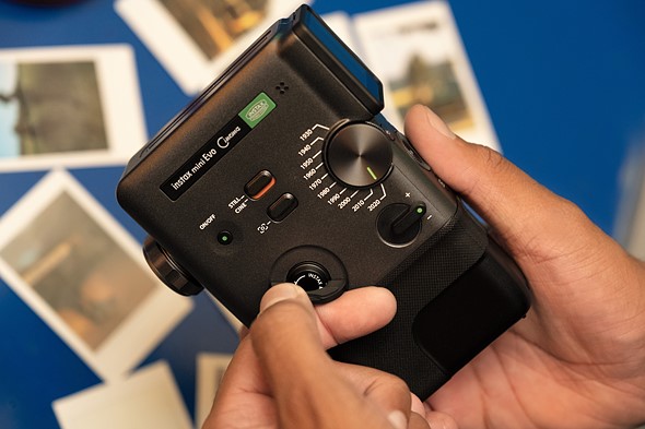 a hand holds a black instax camera over a blue table scattered with instax prints