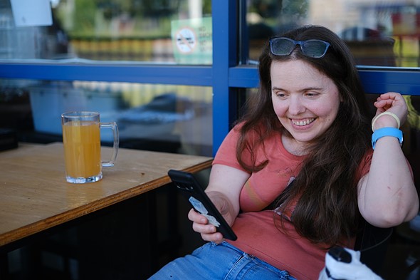 a person sits leaning against windows while smiling and looking at their phone