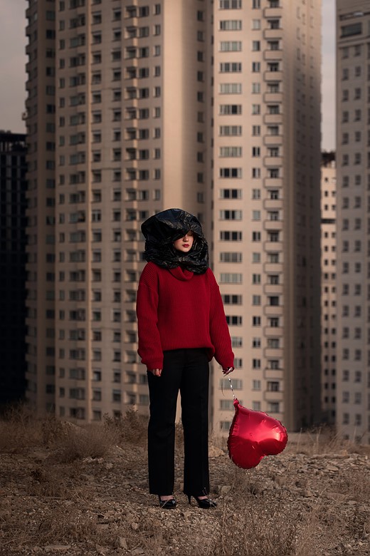 a person wearing a large black hood and red sweater stands holding a sagging red heart baloon in front of skyscrapers
