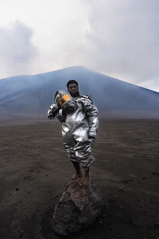 a person wewaring a silver lava protection suit stands on a rock in front of a barren landscape
