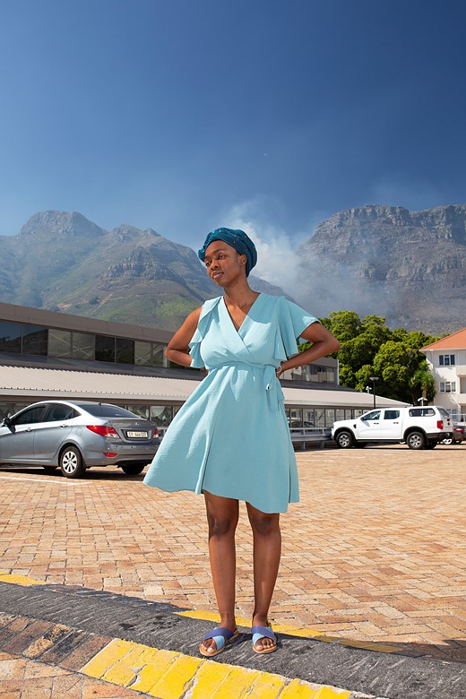 a woman in a blue dress stands in a paring lot with cars houses and mountains behind her