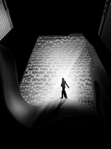 Black and white urban scene of a silhouetted figure standing against a high rise building, showcasing layering and framing in photography with strong architectural contrast.