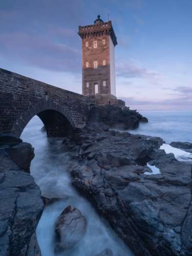 Lighthouse beside a stone bridge at blue hour with long exposure water, illustrating photography framing techniques using natural rock formations and leading lines.