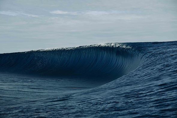 A single large ocean wave curls toward the horizon under a soft overcast sky