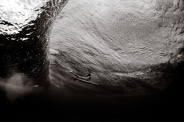 A surfer duck dives under a breaking wave in a black and white underwater scene filled with bubbles and textured water above