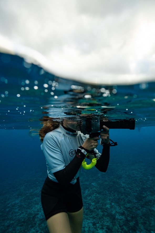 A woman in a rash guard holds an underwater camera housing partially submerged in clear blue ocean water beneath a bright cloudy sky