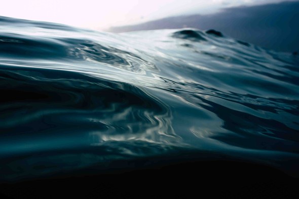 Dark ocean waves roll gently with soft reflections of light on their surface under a bright hazy sky