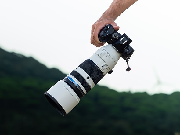 a hand holds a camera with a large white telephoto lens attached in front of a blurred green forest and white sky