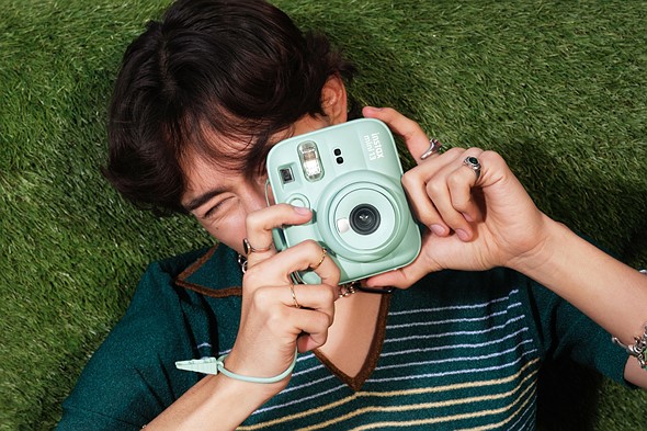 a person wearing a striped shirt lies on green turf while holding a green instax instant camera in front of their face