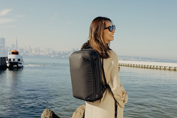 a woman wearing sunglasses and a black backpack stands smiling at a harbor with a city in the background