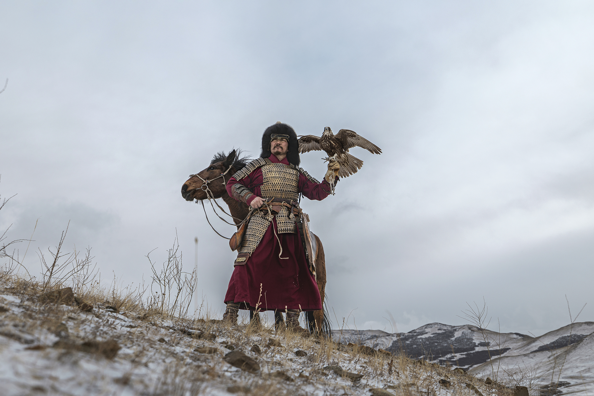 Portrait by Ganzorig Miimaa of a Mongolian eagle hunter in traditional armor and fur hat standing beside his horse, holding a trained falcon against a dramatic cloudy winter steppe sky.